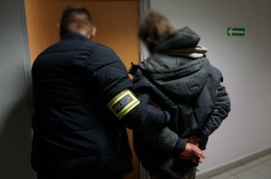 Na zdjęciu nieumundurowany policjant, który ma na ramieniu założoną opaskę odblaskową z napisem Policja prowadzi do jednego z pomieszczeń służbowych na przesłuchanie zatrzymanego mężczyznę zakutego w kajdanki na ręce trzymane z tyłu
