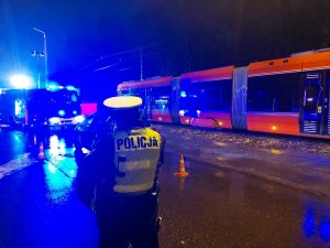 Na zdjęciu widac policjanta z drogówki w żółtej kamizelce odblaskowej podczas prowadzenia czynności na miejscu wypadku drogowego. W tle widac wóz Państwowej Straży Pożarnej oraz Tramwaj uczestniczący w zdarzeniu