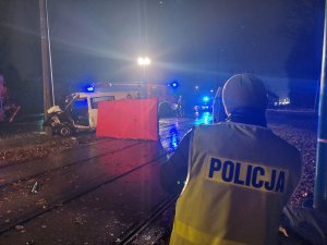 Na zdjęciu widac policjanta z drogówki w żółtej kamizelce odblaskowej podczas prowadzenia czynności na miejscu wypadku drogowego. W tle widac wóz Państwowej Straży Pożarnej oraz volkswagena uczestniczącego w zdarzeniu