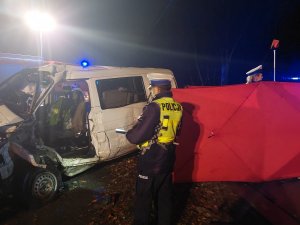 Na zdjęciu widac policjanta z drogówki w żółtej kamizelce odblaskowej podczas prowadzenia czynności na miejscu wypadku drogowego. W tle widac uszkodzonego volkswagena uczestniczącego w zdarzeniu