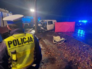 Na zdjęciu widac policjantów z drogówki w żółtej kamizelce odblaskowej podczas prowadzenia czynności na miejscu wypadku drogowego. W tle widac wózy Państwowej Straży Pożarnej oraz uszkodzonego volkswagena uczestniczącego w zdarzeniu