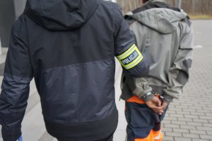 Na zdjęciu nieumundurowany policjant z opaską odblaskową na ramieniu prowadzi do świętochłowickiej komendy zatrzymanego mężczyznę zakutego w kajdanki na ręce trzymane z tyłu