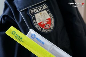 Na zdjęciu dwie opaski odblaskowe w kolorze białym oraz żółtym z logiem Policji. Za nimi w tle framgment granatowej kurtki służbowej z naszywką przedstawiająca orła oraz symbol ruchu drogowego na tle polskiej flagi oraz napis Wydział Ruchu Drogowego wokół. Na górze naszywki widać napis Policja