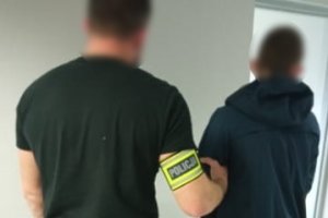 Na zdjęciu nieumundurowany policjant z założoną na prawym ramieniu opaską osblaskową z napisem Policja oraz zatrzymany mężczyzna zakuty w kajdanki na ręce trzymane z przodu