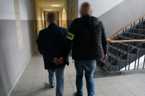 Na zdjęciu nieumundurowany policjant z opaską odblaskową z napisem Policja na ramieniu prowadzi wewnątrz budynku świętochłowickiej komendy zatrzymanego mężczyznę zakutego w kajdanki na ręce trzymane z tyłu