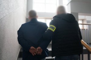 Na zdjęciu nieumundurowany policjant z opaską odblaskową z napisem Policja na ramieniu prowadzi wewnątrz budynku świętochłowickiej komendy zatrzymanego mężczyznę zakutego w kajdanki na ręce trzymane z tyłu