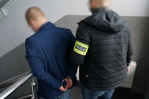 Na zdjęciu nieumundurowany policjant z opaską odblaskową z napisem Policja na ramieniu prowadzi wewnątrz budynku świętochłowickiej komendy zatrzymanego mężczyznę zakutego w kajdanki na ręce trzymane z tyłu