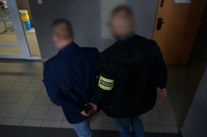 Na zdjęciu nieumundurowany policjant z opaską odblaskową z napisem Policja na ramieniu prowadzi wewnątrz budynku świętochłowickiej komendy zatrzymanego mężczyznę zakutego w kajdanki na ręce trzymane z tyłu