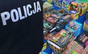 Na zdjęciu zbliżenie na napis Policja na kurtce umundurowanego policjanta, po prawej stronie widać róznego rodzaju materiały piroteczniczne wystawione na stoisku