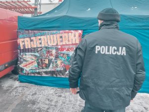 Na zdjęciu policjant dzielnicowy stojący twarzą do namiotu w którym sprzedawane są fajerwerki. Na namiocie widac plakat z napisem fajerwerki oraz grafiką wyrobów pirotechnicznych