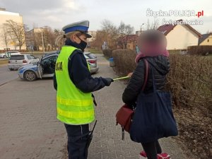 Na zdjeciu mundurowa z drogówki w trakcie wręczania pieszej opaski odblaskowej. W tle widać policyjny radiowóz