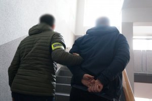 Na zdjęciu nieumundurowany policjant, który ma założoną na prawym ramieniu opaskę odblaskową z napisem Policja prowadzi wewnątrz budynku świętochłowickiej komendy zatrzymanego mężczyznę zakutego w kajdanki na ręce trzymane z tyłu. Przemieszczają się schodami na wyższą kondygnację w budynku.