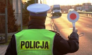 Na zdjęciu policjant z drogowki który trzyma w prawej dłoni tzw. lizak, służący do zatrzymywania pojazdów oraz wskazywania kierującym miejsca postoju. W tle widać zbliżający się pojazd.