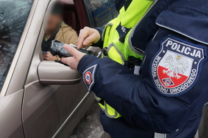 Na zdjęciu zbliżenie na policjanta który na specjalnym przenośnym urzadzeniu sprawdza w policyjnych bazach danych zatrzymanego do kontroli drogowej kierowcę. W tle widac mężczyznę oczekującego na sprawdzenie wewnątrz pojazdu.