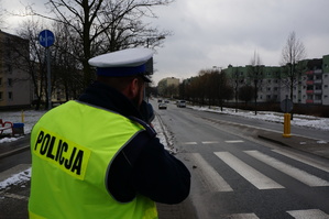 Na zdjęciu policjant z drogowki ubrany w kamizelkę odblaskową z napisem Policja w okolicy przejścia dla pieszych w trakcie dokonywania pomiaru prędkości zbliżającego się pojazdu, widocznego w tle.