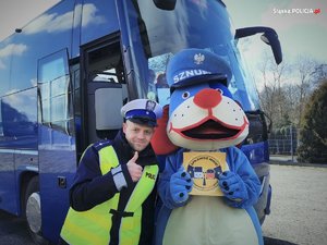 Na zdjęciu policjant z drogówki wraz z maskotką śląskiej Policji psem Sznupkiem na tle autokaru