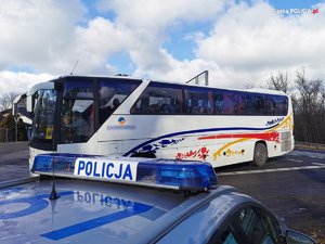 Na zdjęciu zbliżenie na napis Policja na belce oświetleniowej radiowozu, w tle widać autobus z młodzieżą