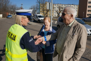 Na zdjęciu mundurowa przy jednym ze świętochłowickich przejść dla pieszych. W tle widać pojazdy które zatrzymały się przed przejściem dla pieszych oraz policyjny radiowóz. Policjantka zakłada na rękę seniora opaskę odblaskową. Obok mężczyzny stoi starsza kobieta.