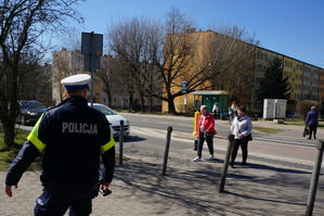 Na zdjęciu mundurowy w trakcie prowadzenia obserwacji zachowania kierujących wobec pieszych w okolicy jednego z przejść. W tle widać przechodzace przez pasy dwie seniorki.