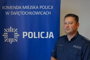 Na zdjęciu dzielnicowy aspirant Rafał Gabryś, który stoi na tle granatowej ścianki z napisem Komenda Miejska Policji w Świętochłowicach oraz logiem formacji z napisem Policja