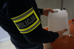 Na zdjęciu zbliżenie na napis policja na opasce, którą nieumundurowany policjant ma założoną na ramieniu. Policjant trzyma butelkę z zabezpieczonym trunkiem niewiadomego pochodzenia.