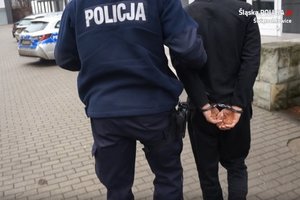 Na zdjęciu umundurowany policjant prowadzi do budynku świętochłowickiej komendy zatrzymanego mężczyznę zakutego w kajdanki na ręce trzymane z tyłu