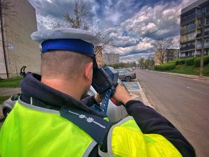 na zdjęciu na terenie otwartym umundurowany policjant w białej czapce służbowej, w kamizelce odblaskowej, w ręce trzyma urządzenie laserowe do mierzenia prędkości, które kieruje w stronę ulicy