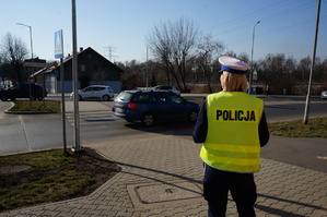 na zdjęciu umundurowana policjantka w odblaskowej kamizelce z napisem policja. Stoi przed skrzyżowaniem