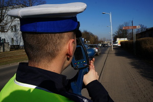 na zdjęciu umundurowany policjant w odblaskowej kamizelce z napisem policja. w ręce trzyma urządzenie do meirzenia prędkości, skierowane na ulicę