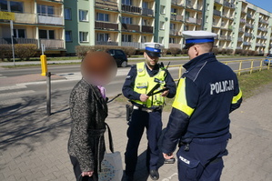 na zdjęciu dwóch policjantów umundorowanych, w kamizelce odblaskowej z napisem policja. Przy nich stoi kobieta- przechodizłą przez przejście dla pieszych