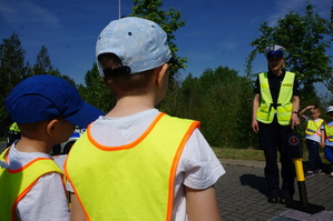 Na zdjeciu widac dzieci oraz policjantkę która objasnia im zasady przechodzenia przez jezdnię oraz znaczenie sygnalizacji świetlnej.