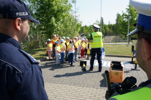 Zdjecie zrobione zza pleców dwóch mundurowych. W tle widać policjantkę oraz dzieci. Mundurowa tłumaczy im zasady bezpieczeństwa na drodze