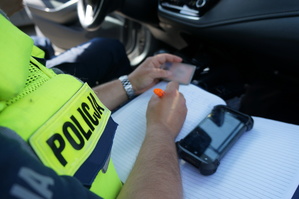 Zbliżenie na napis policja na kamizelce odblaskowej policjanta z drogówki, który siedzi w radiowozie i dokonuje sprawdzenia w policyjnych systemach zatrzymanego do kontroli drogowej kierującego.