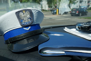 Na zdjęciu wewnątrz radiowozu czapka służbowa policjanta drogówki z białym otokiem i orłem na przodzie oraz teczka z symbolem Ruchu Drogowego. W tle widać przejście dla pieszych i przemieszczające się pojazdy.