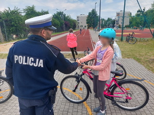 Na zdjęciu widać policjanta który gratuluje jednej z dziewczynek zdanego egzaminu na kartę rowerową. W tle widać dzieci przebywające na boisku szkolnym