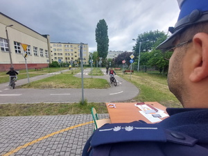 zbliżenie na pagon na ramieniu policjanta który czuwa nad przebiegiem egzaminu na karte rowerową. W tle widać uczestniczki pokonujące specjalny tor znajdujący się na szkolnym miasteczku ruchu drogowego