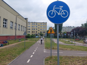 Na pierwszym planie znak informujący o drodze dla rowerów. W tle widać uczestniczki egzaminu na kartę rowerową, pokonujące tor na terenie Miasteczka Ruchu Drogowego