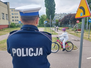 Policjant w trakcie czuwania nad przebiegiem egzaminu na karte rowerową. Na zdjęciu widać jak młode osoby poruszające się na rowerach pokonują tor na terenie Miasteczka Ruchu Drogowego