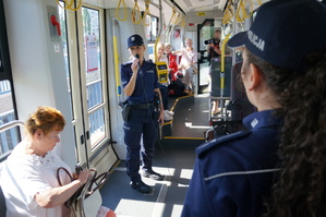 Wnętrze tramwaju, w ktorym widać dwie policjantki oraz seniorów. Policjantka trzyma mikrofon w dłoni i prowadzi prelekcję dla uczestników akcji profilaktycznej