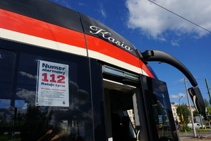Zbliżenie na napis Kasia na przodzie czerwonego tramwaju. Na szybie tramwaju widać plakat informujący o numerze alarmowym 112