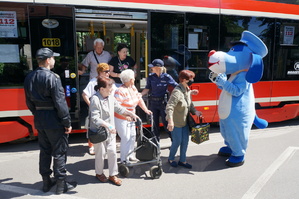 Na zdjęciu widac seniorów wychodzących z tramwaju. Wita ich policjantka i strażak oraz maskotka śląskiej Policji - pies Sznupek