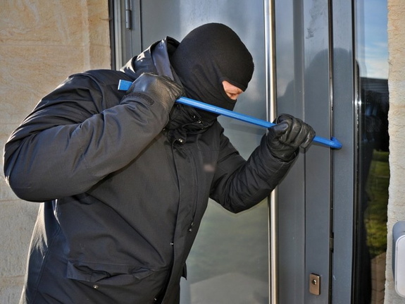 Na zdjęciu mężczyzna w kominiarce na głowie, który próbuje otworzyć drzwi za pomocą metalowego przedmiotu, który trzyma w dłoni