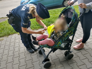 Policjantka umundurowana stoi przy kobiecie, która prowadzi wózek z dzieckiem. Policjantka nachyla się nad dzieckiem