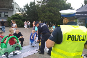 na zdjeciu dzieci przechodzące przez mini miasteczko ruchu drogowego, Policjant umundurowany w tle