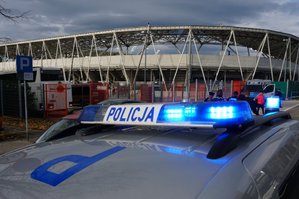 Zbliżenie na napis policja na belce oświetleniowej. W tle widać stadion