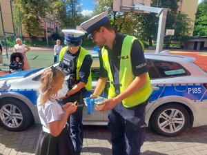 Na zdjęciu policjanci wraz z dziewczynką która ogląda otrzymany od nich policyjny odblask. Jeden z policjantów trzyma koszyczek z odblaskami i słodyczami