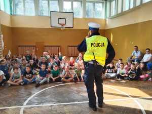 Na zdjęciu mundurowy stojący plecami do obiektywu oraz siedzące przed nim dzieci. Policjant opowiada im o zasadach którymi muszą się kierować aby droga do szkoły była dla nich bezpieczna.