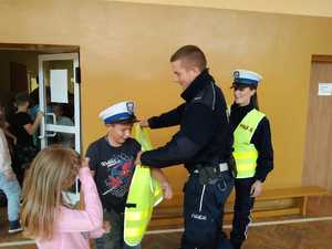 Na zdjęciu mundurowy pomaga założyć dziecku kamizelkę odblaskową z napisem policja. Obok stoi umundurowana policjantka.