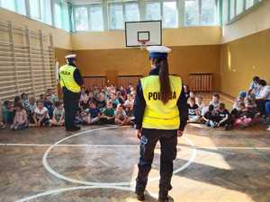 Na zdjęciu mundurowi stojący plecami do obiektywu oraz siedzące przed nimi dzieci. Policjanci opowiadają im o zasadach którymi muszą się kierować aby droga do szkoły była dla nich bezpieczna.