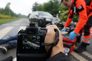 Na zdjęciu zbliżenie na wyświetlacz aparatu fotograficznego na którym widac symylację wypadku drogowego. W tle widac uczestników scenki.